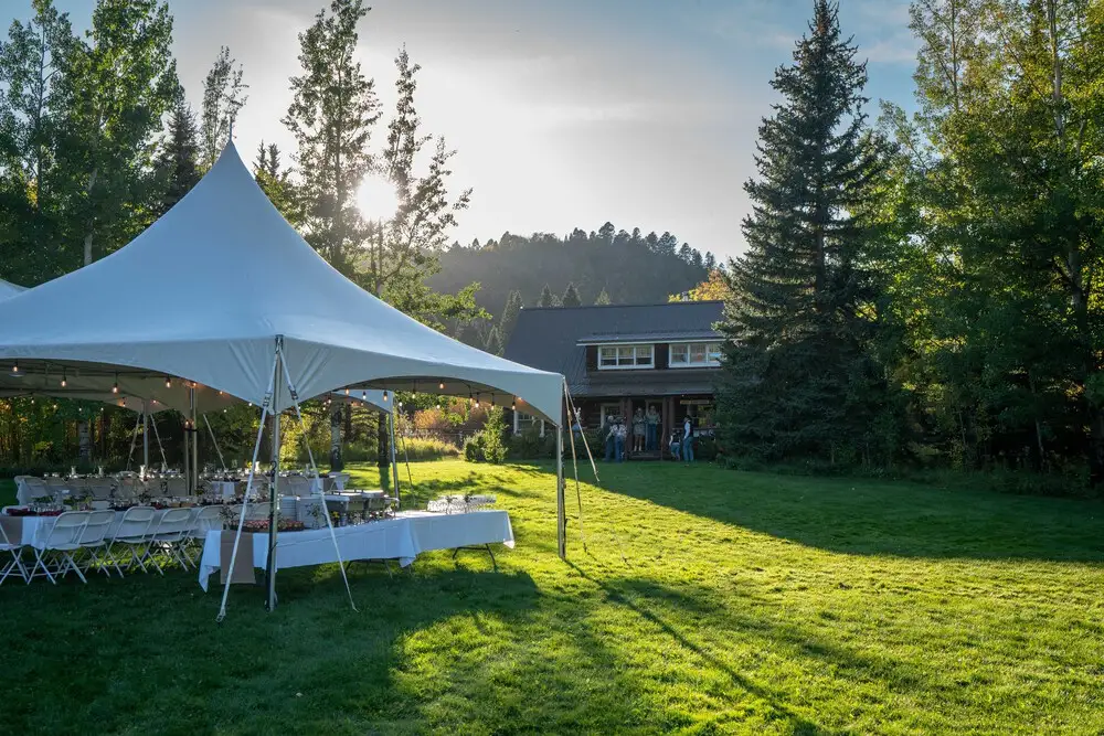 A white tent in a grassy field with a table set for a meal. People are standing around the tent, enjoying the outdoor gathering