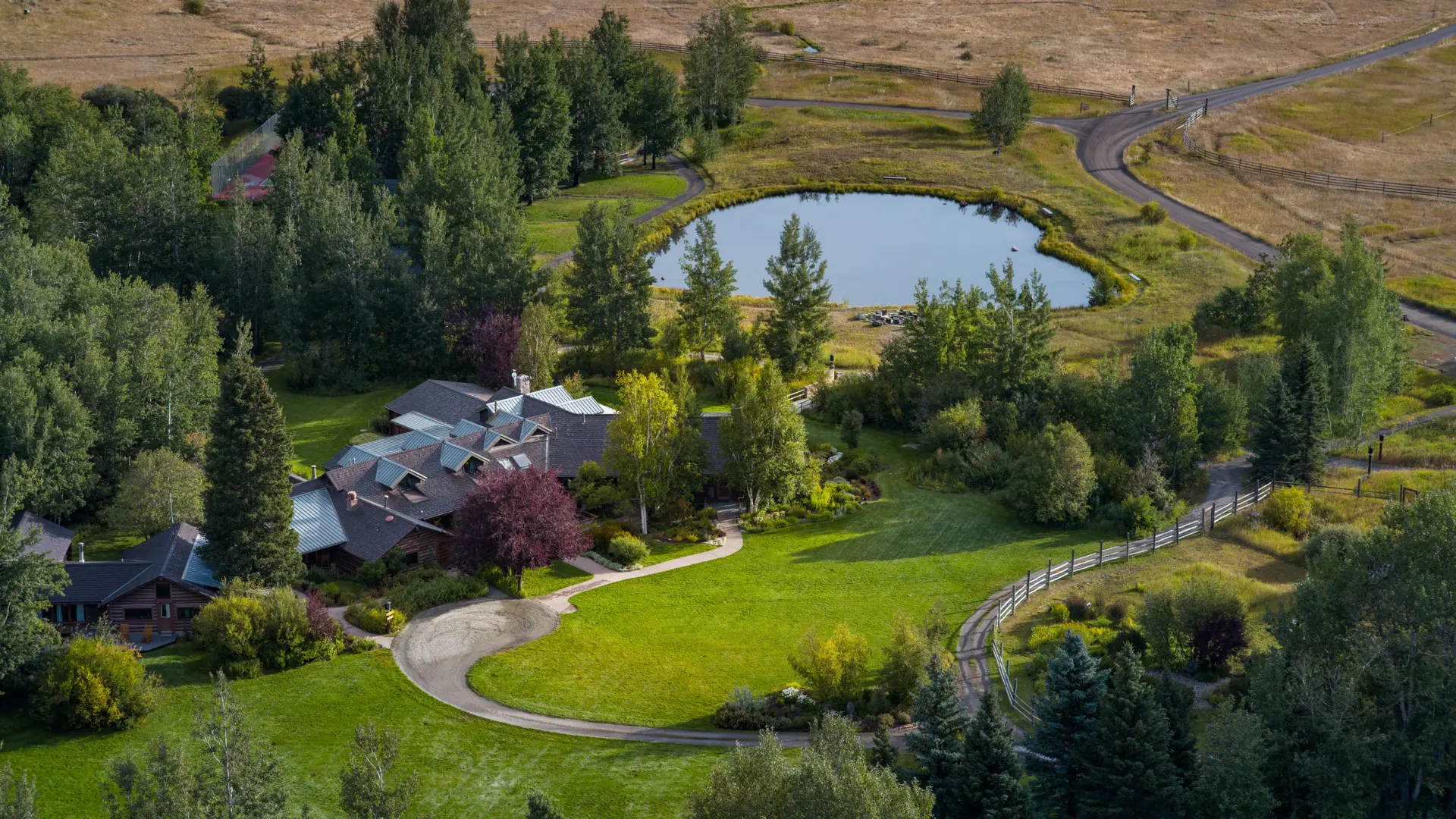 A large house with a circular driveway and a pond in the middle of the yard. The house is surrounded by trees and has a green roof. The driveway is curved and leads to the house. The pond is in the center of the yard and is surrounded by trees and bushes