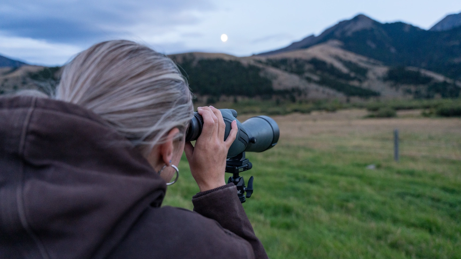 A woman looking at the moon through binoculars in a field with mountains in the background