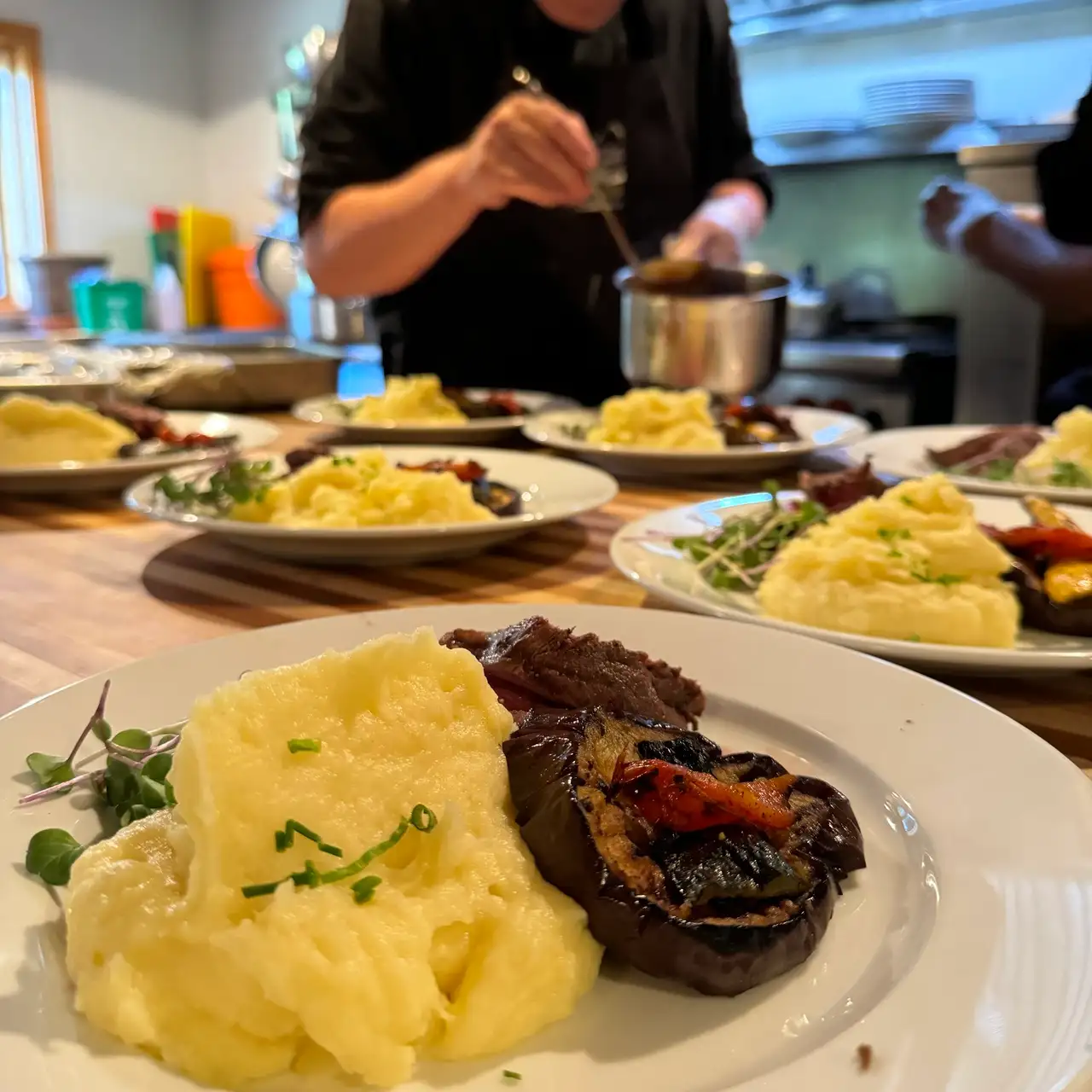 A person is serving food on a table with several plates of food. The food includes mashed potatoes, meat, and vegetables. The person is scooping the food from a pot onto the plates