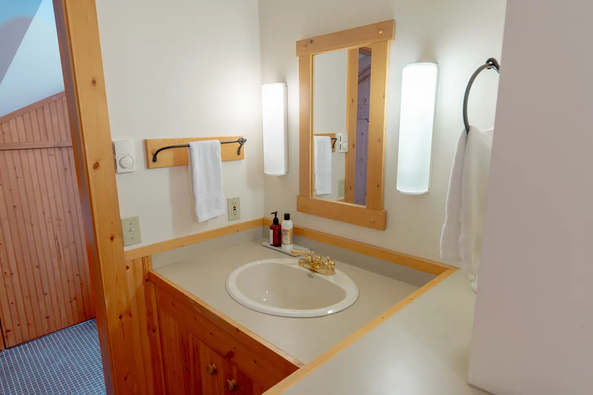 A bathroom with a sink, mirror, and towel rack. The sink is white and the mirror is above it. There are two towel racks, one on the left and one on the right. A bottle is placed on the sink. The bathroom has a wooden theme and is well lit