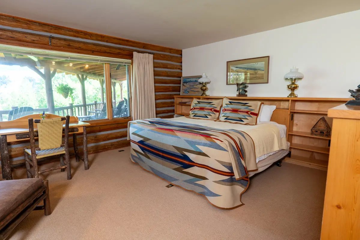 A bedroom with a cozy bed, a chair, a table, and a window. The room has a rustic feel with wooden walls and a log cabin design