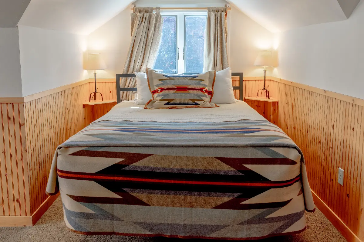 A bedroom with a large bed, a window, and a lamp. The bed has a striped bedspread and a pillow with a red and white design.