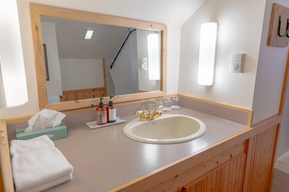 A bathroom with a sink and mirror. The sink is white and the counter is made of wood. There is a large mirror above the sink. Above the mirror, there is a light. The bathroom has a clean and bright appearance