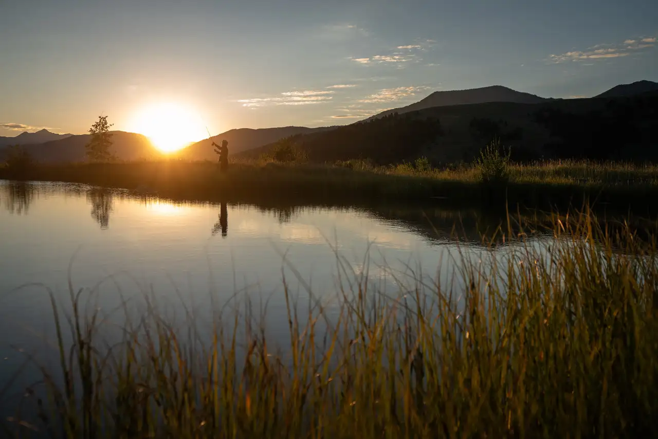 A person standing on a hillside, looking down at a body of water, with the sun setting in the background