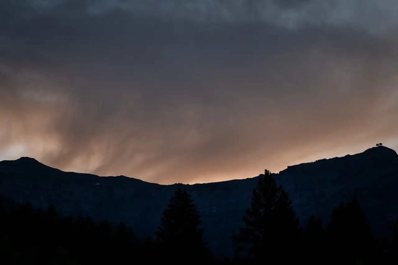 A dark mountain with a cloudy sky above it, with a few trees in the foreground and a sunset in the background