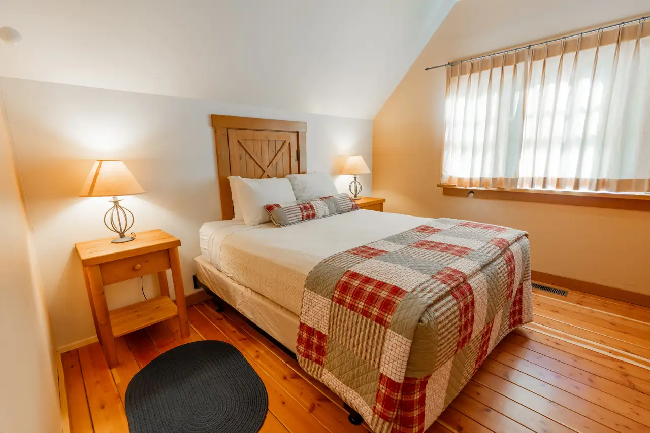 A bedroom with a bed under a window, a plaid blanket, and two lamps on either side