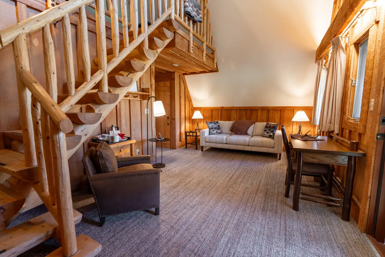 A cozy living room with wooden walls and floor, furnished with a couch, chair, dining table, and staircase