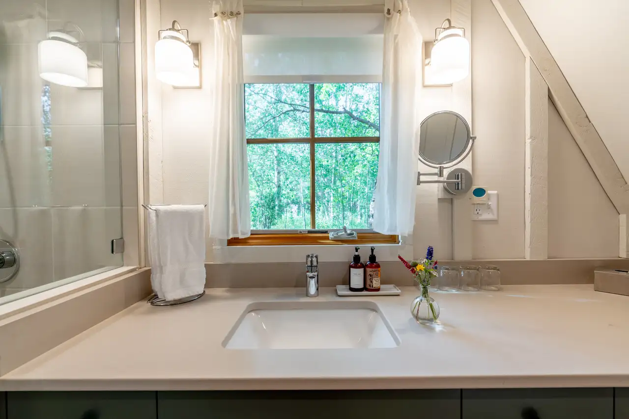 A bathroom with a sink, mirror, and window. The sink is white and the countertop is also white. There are two bottles on the counter, one of which is red. A vase with flowers is also present on the counter