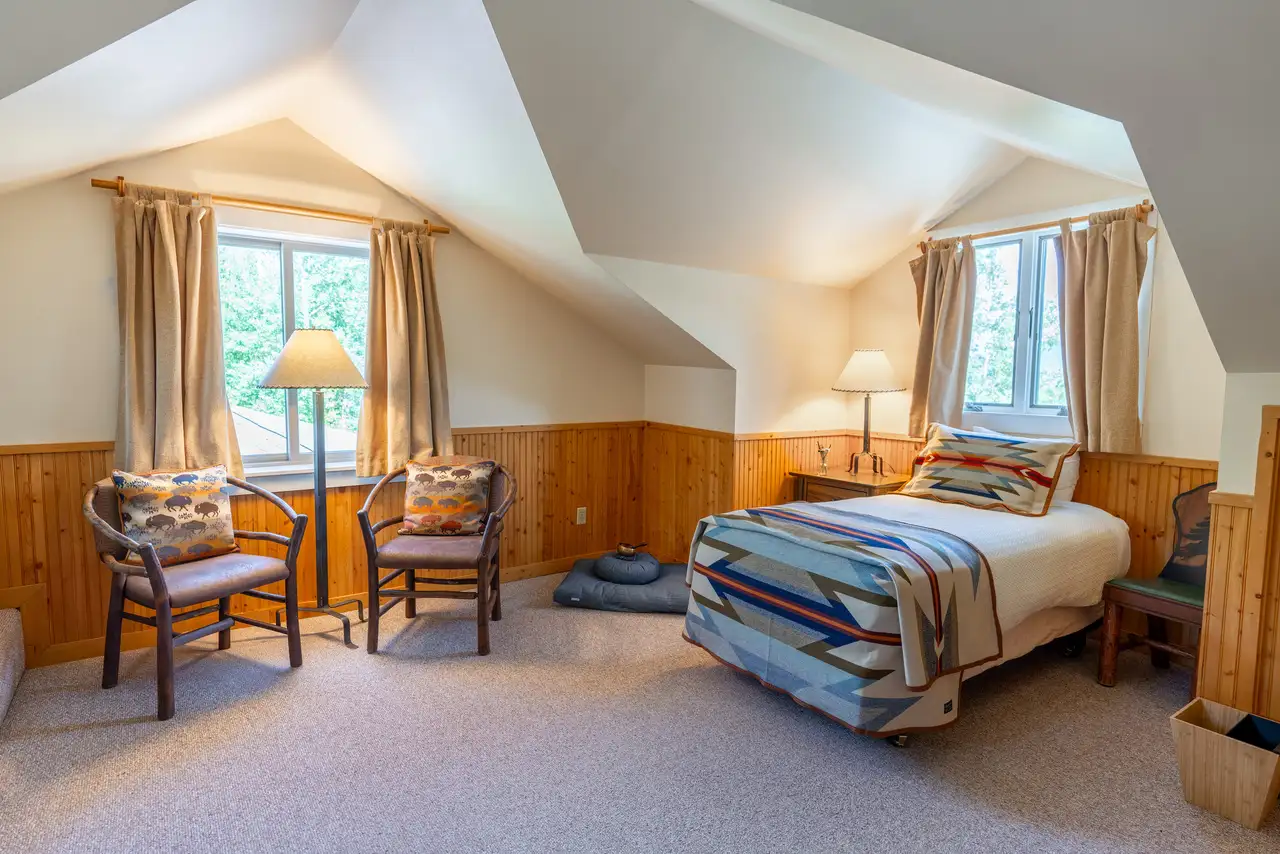 A bedroom with a bed, two chairs, and a window. The bed is covered with a striped comforter and there is a window seat nearby