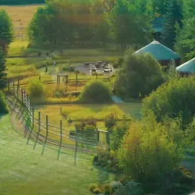 A large grassy field with a fence and a gate, surrounded by a forest. The field has several picnic tables, benches, and chairs, as well as a few houses and a dome-shaped building in the background