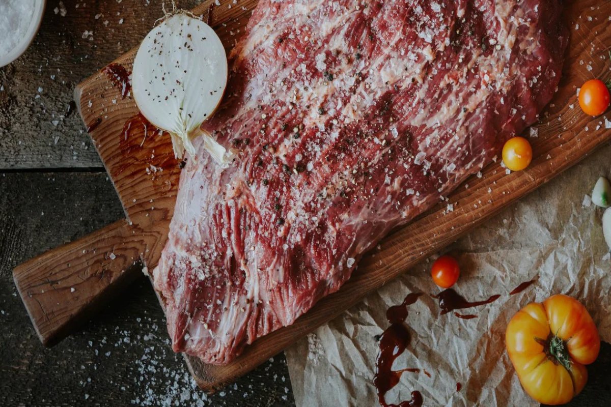 A piece of meat on a cutting board with tomatoes and seasoning