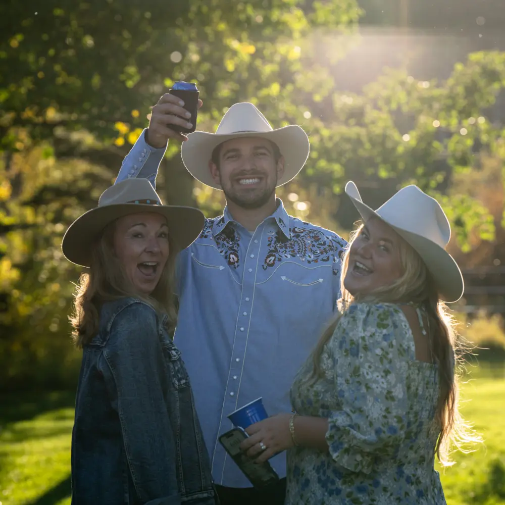 Three people wearing cowboy hats are standing together in a field, smiling and enjoying their time