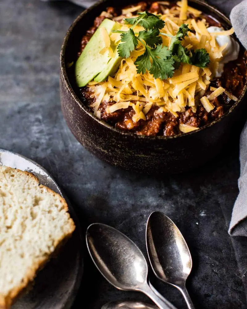 A bowl of chili with cheese and avocado on top, accompanied by a spoon and a piece of bread