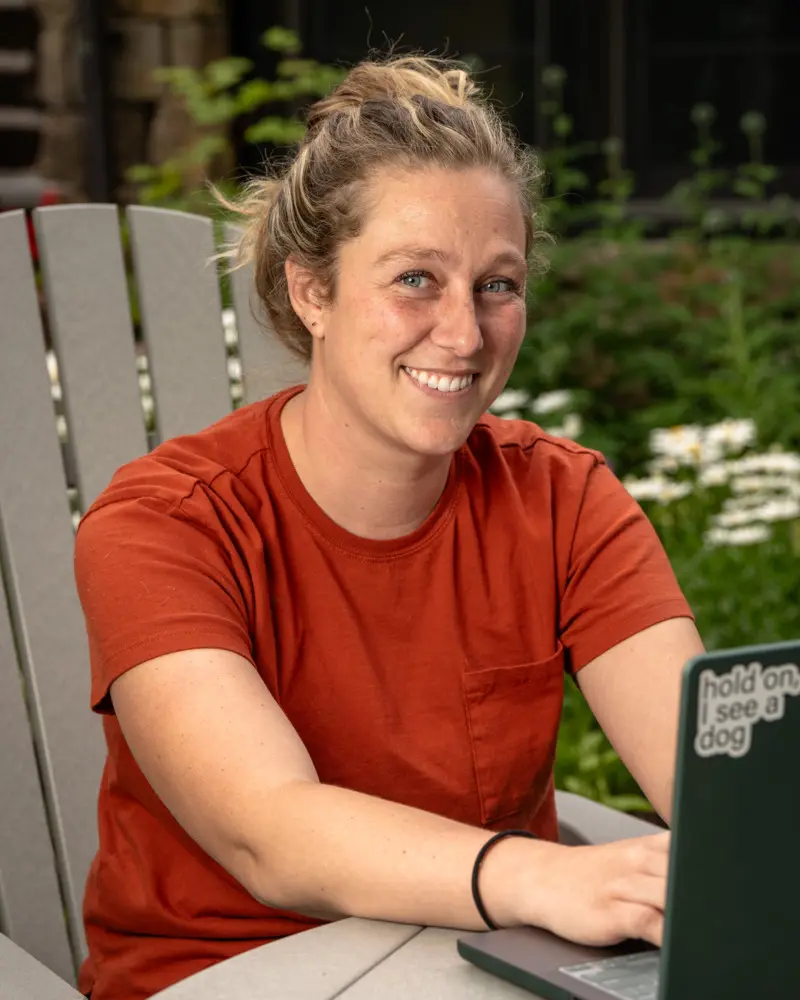 A woman in an orange shirt is smiling while using a laptop