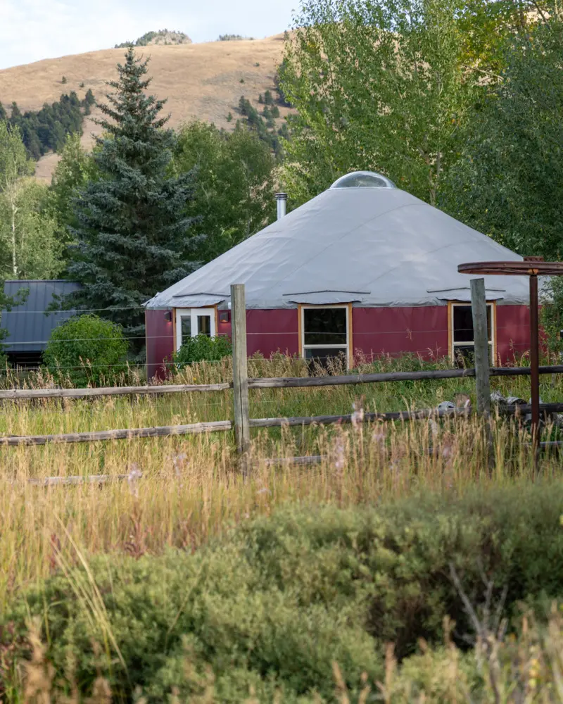 A round house with a red roof and a white dome top. The house is surrounded by a fence and is located in a field with a hill in the background. The house has a chimney and a window on the side