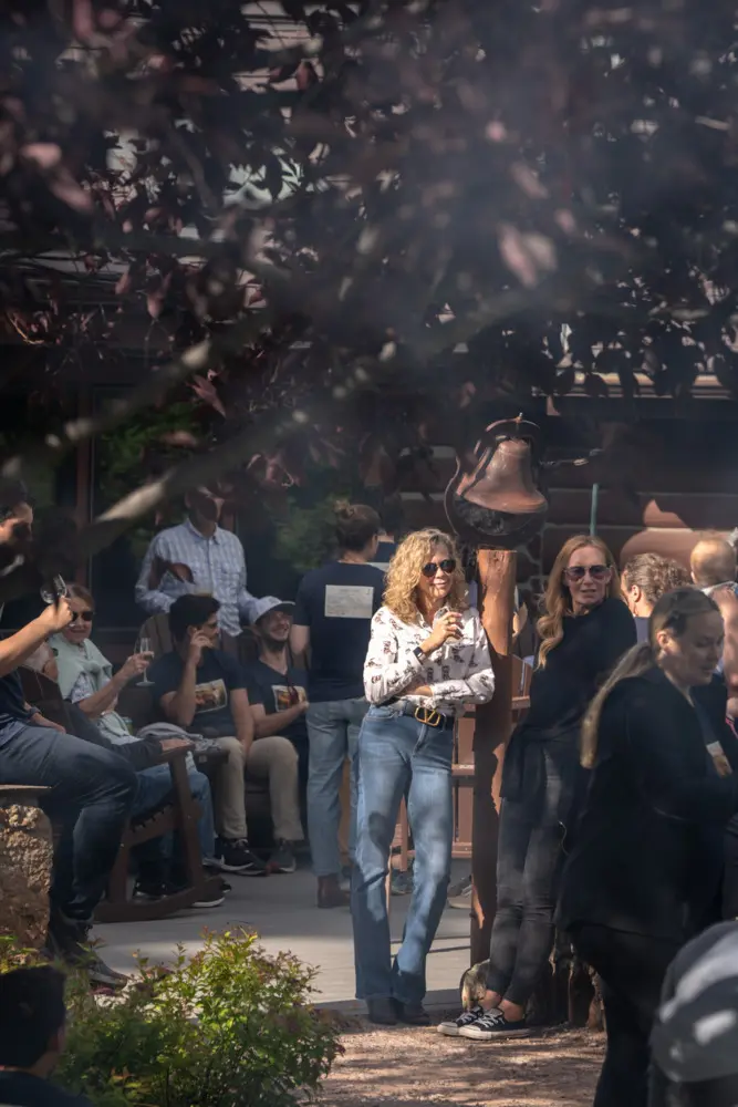 A woman in a white shirt is standing outside, holding a drink and looking at the camera. She is surrounded by a group of people, some of whom are sitting on a bench