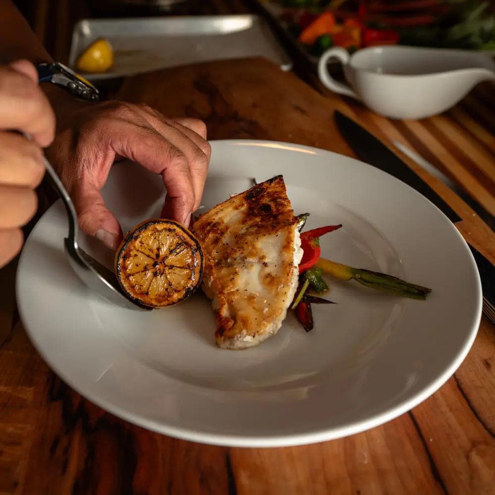 A person cutting fish on a white plate with a slice of lemon and vegetables