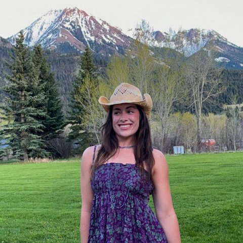 Smiling person in a straw cowboy hat and purple floral sundress standing on a green lawn with snow-capped mountains behind.