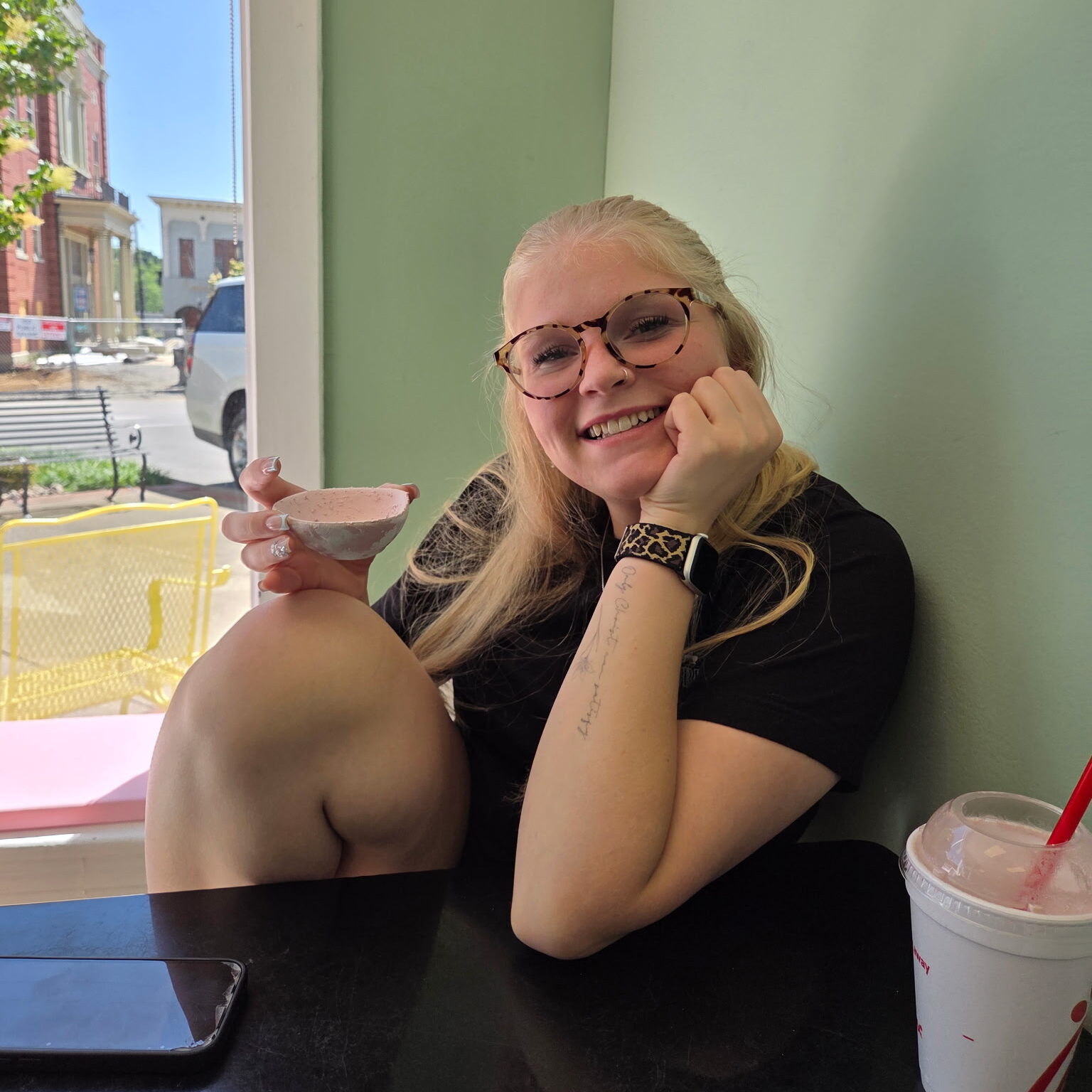 Smiling blonde person in glasses holding a small bowl of pink ice cream at a cafe window table.