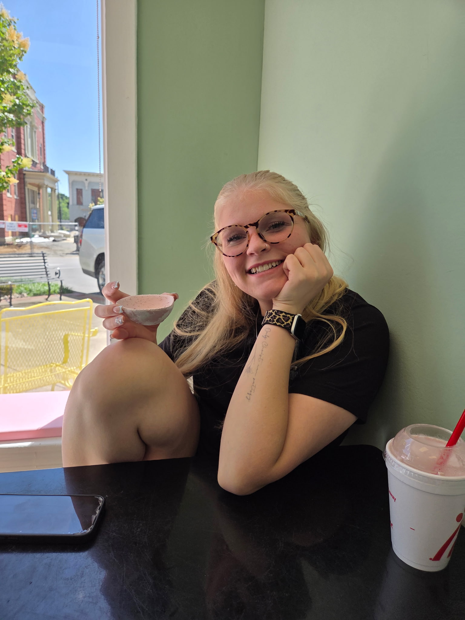 Smiling blonde person in glasses holding a small bowl of pink ice cream at a cafe window table.