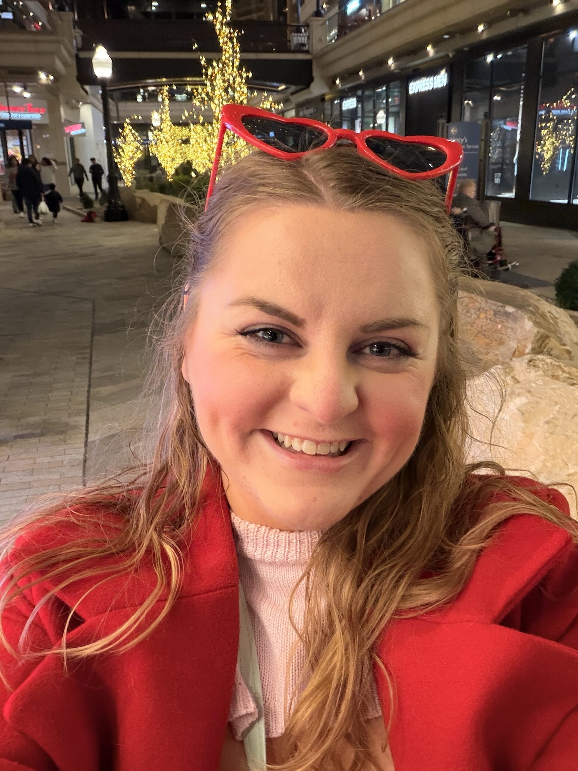Smiling person in a red coat with red heart-shaped sunglasses on their head, taking a selfie at an outdoor shopping center lit with holiday lights.