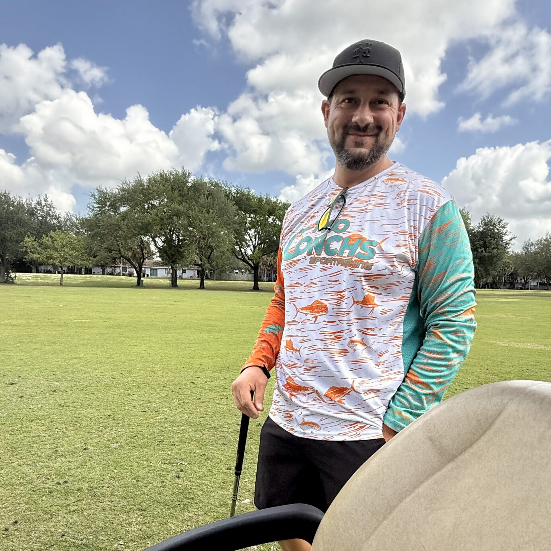 Smiling bearded man in a cap and orange fish-print long-sleeve shirt holding a golf club beside a cart on a sunny course.