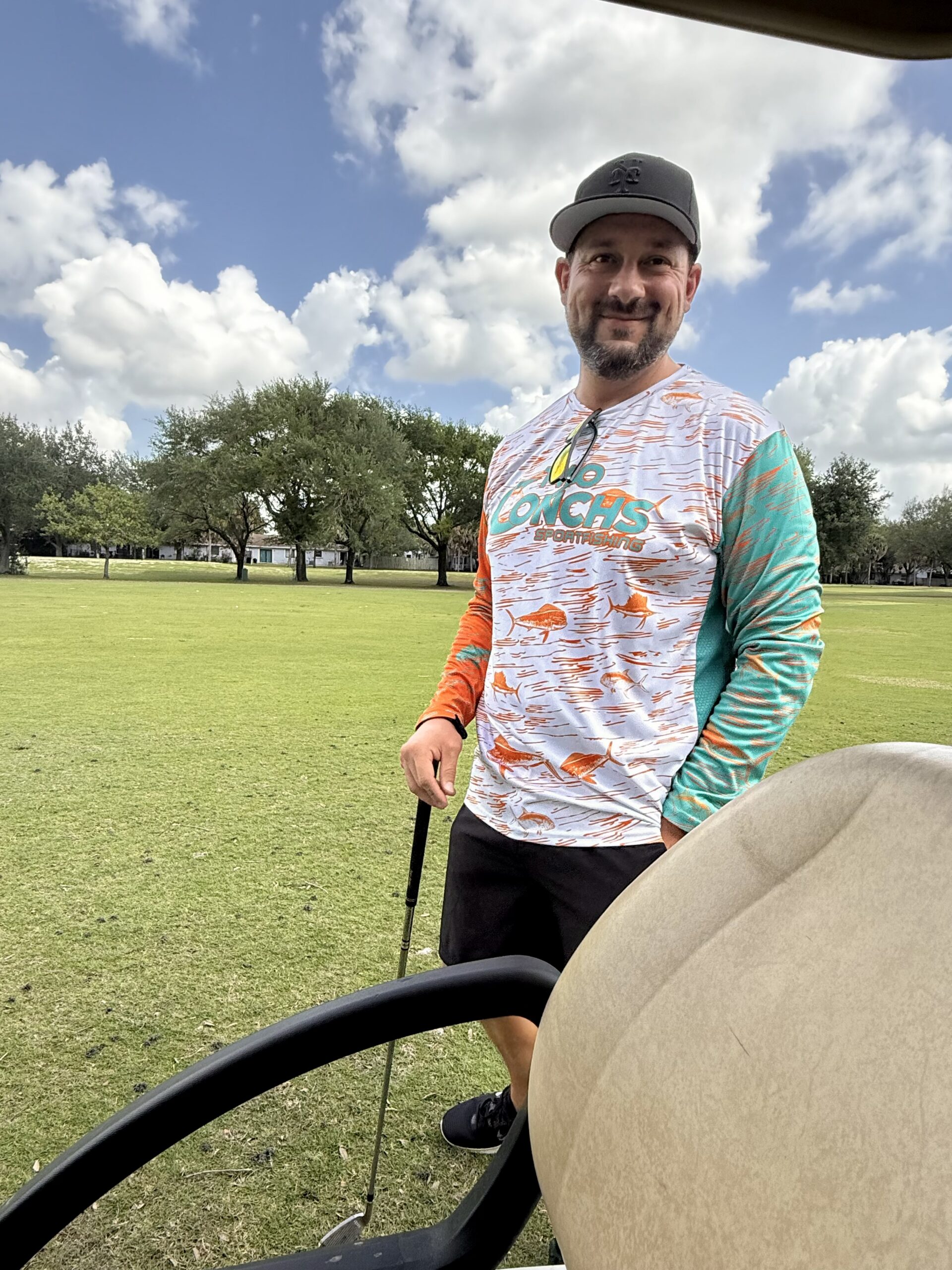 Smiling bearded man in a cap and orange fish-print long-sleeve shirt holding a golf club beside a cart on a sunny course.