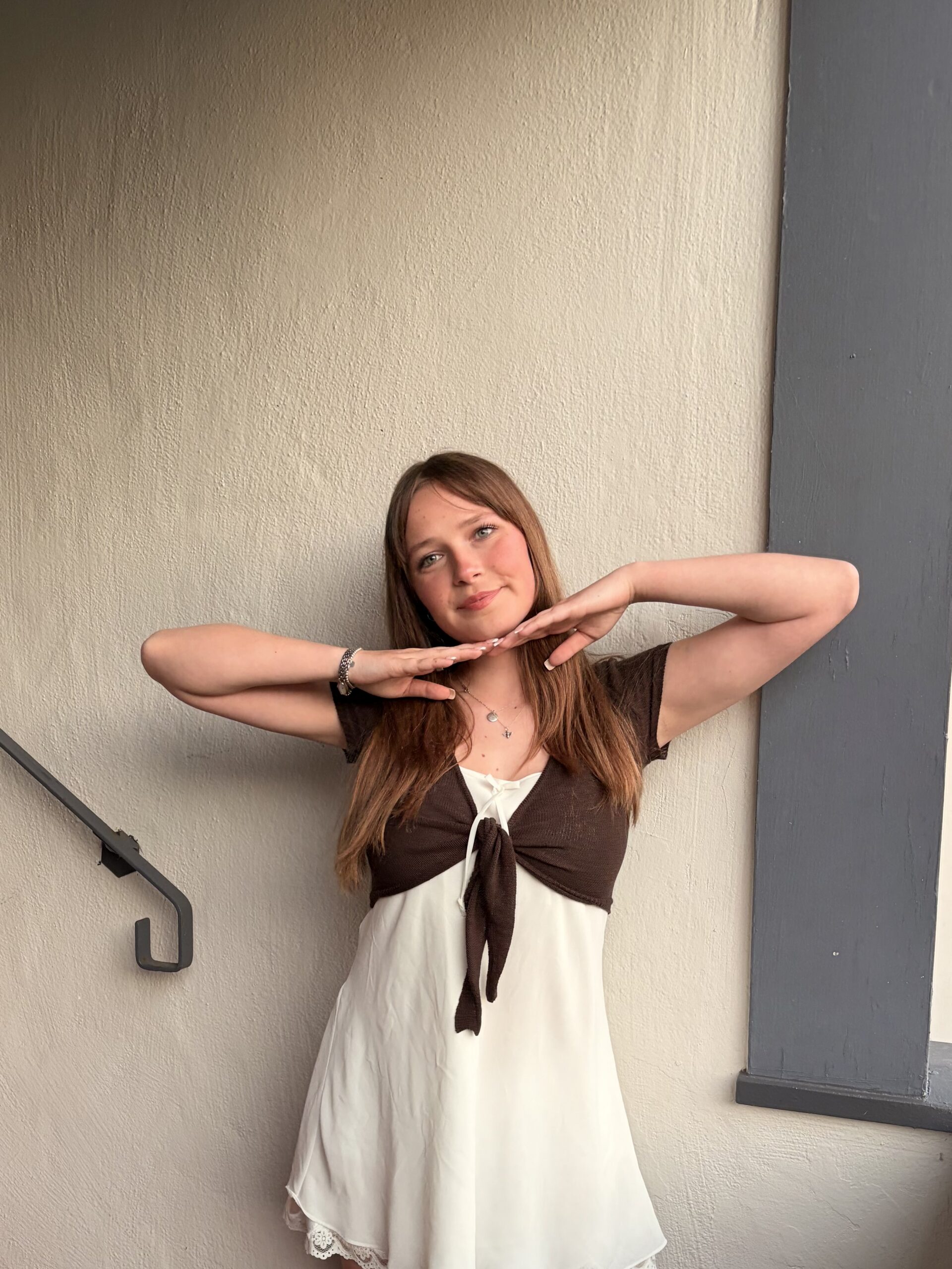 Young woman in a white slip dress and brown tie-front top posing with hands framed under her chin against a beige stucco wall.