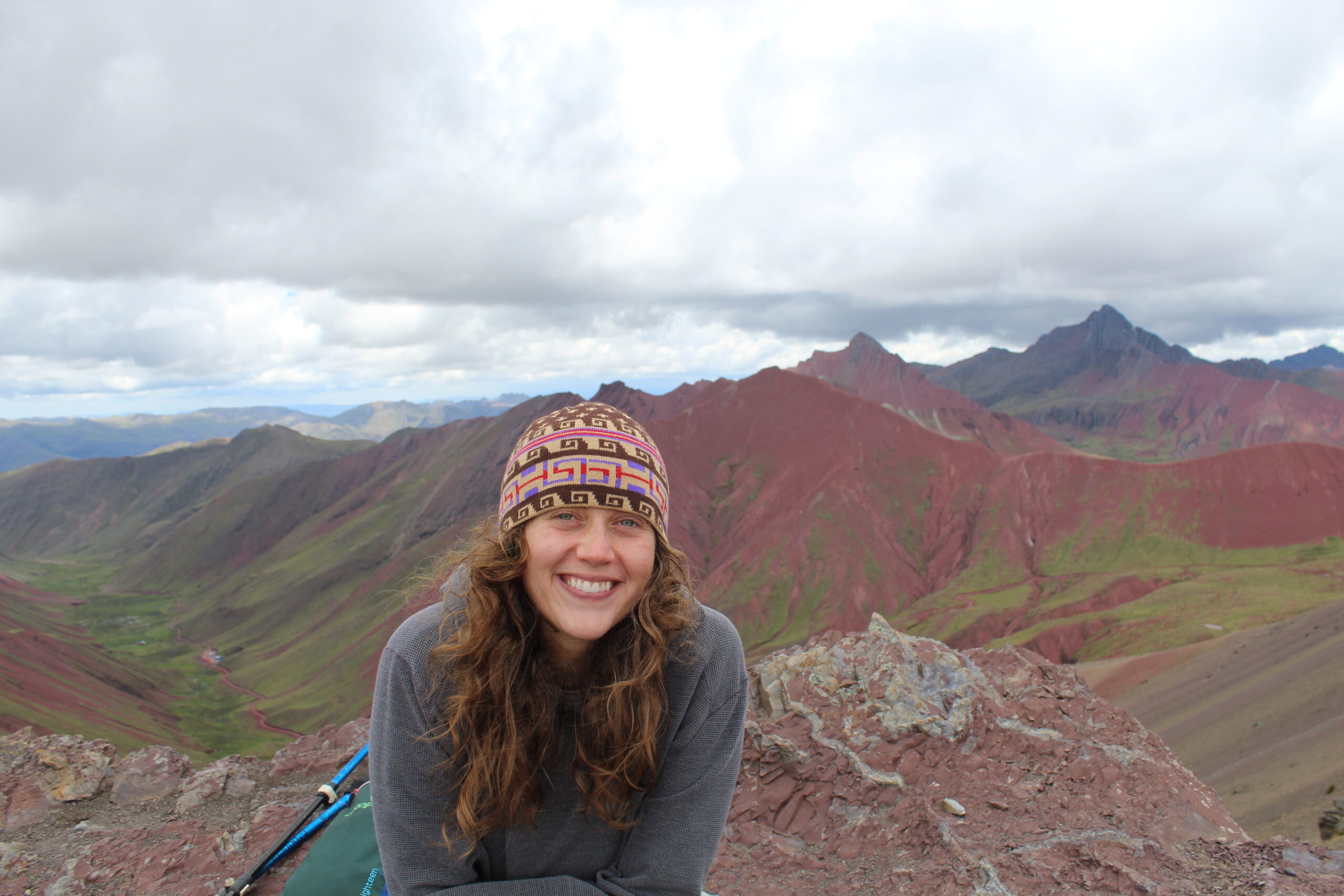 Smiling hiker in a patterned knit beanie on a rocky summit, with red and green Andean mountain ridges behind.