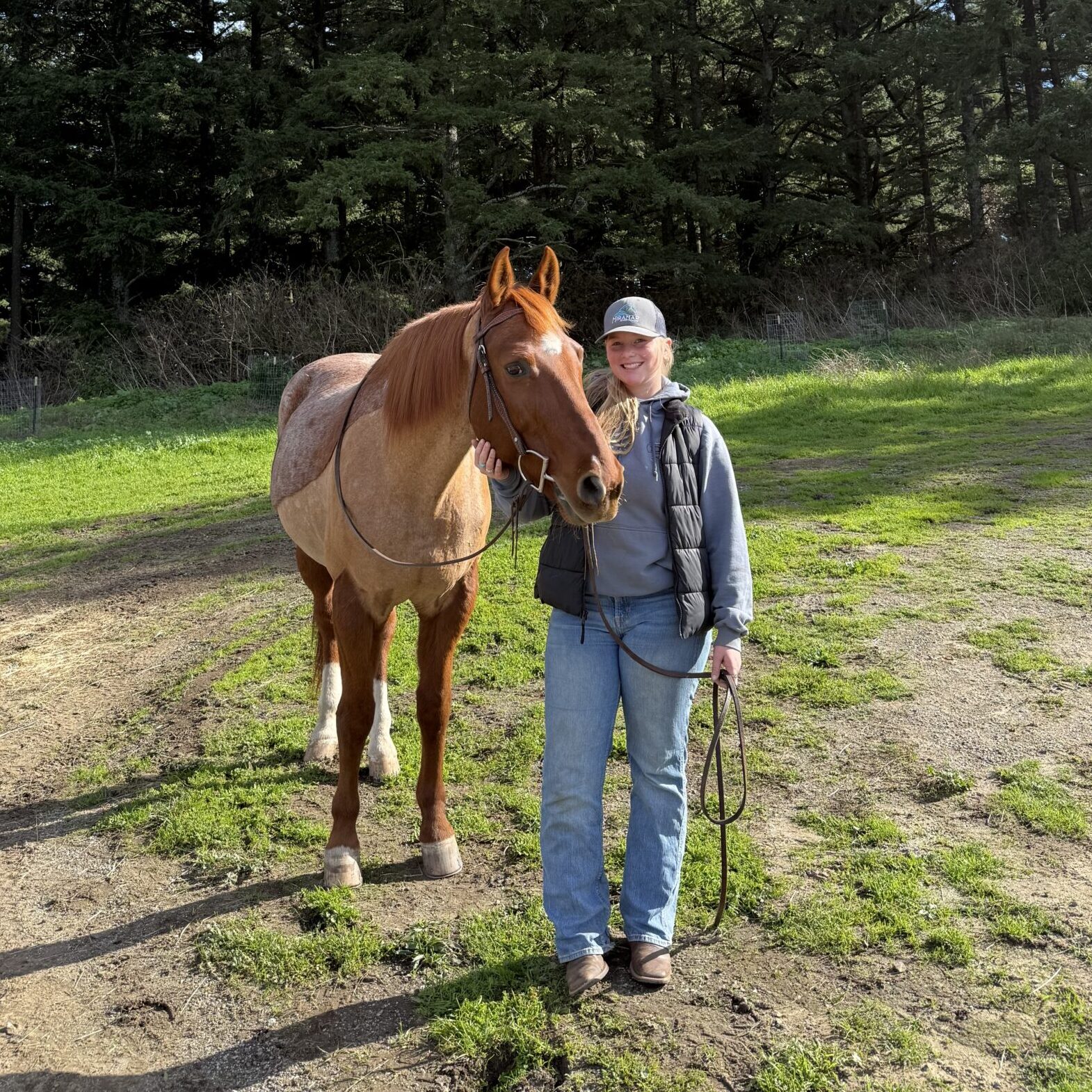 Smiling person in a cap and puffy vest standing beside a chestnut roan horse in a grassy field edged by tall evergreens.