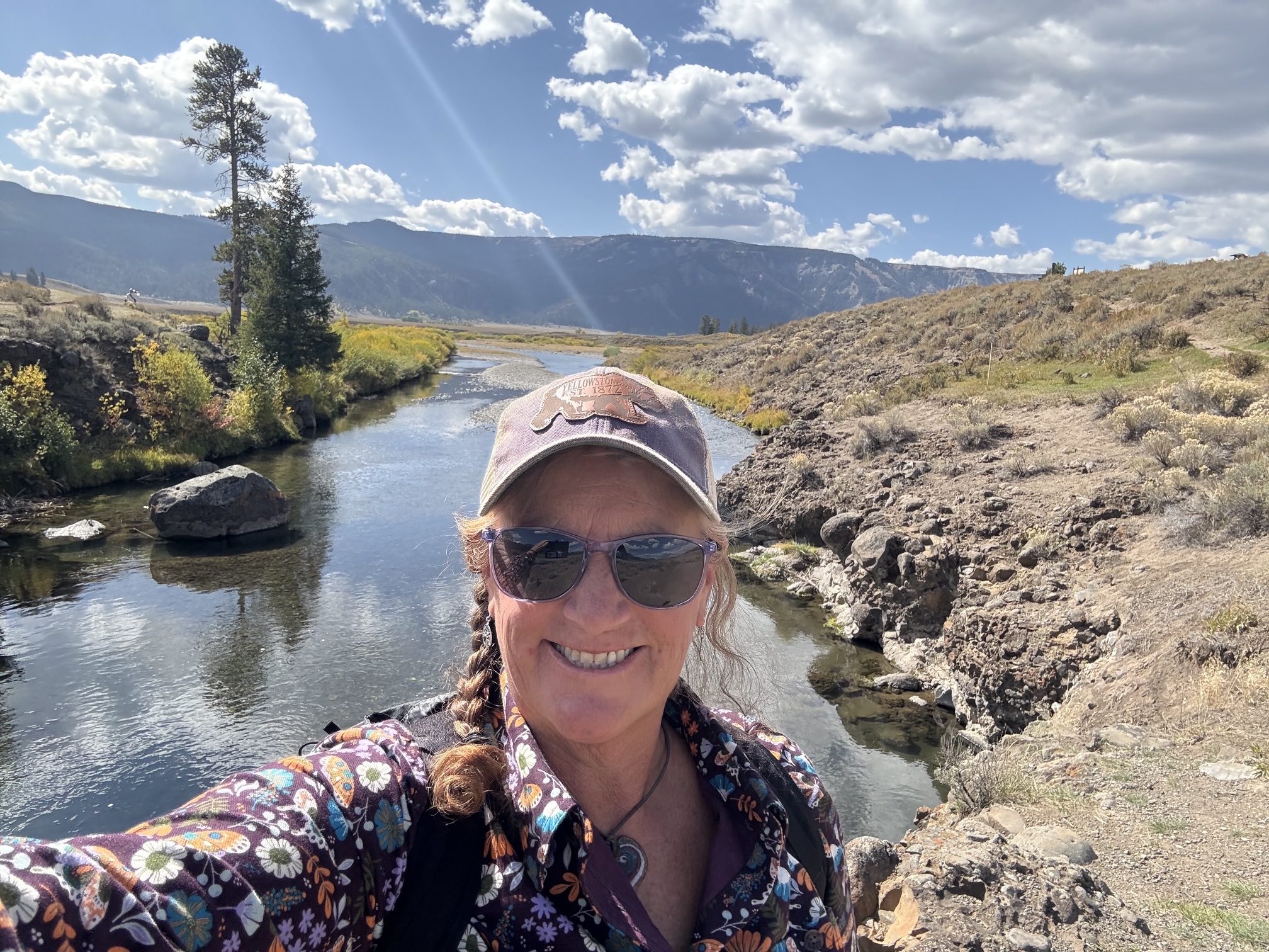 Smiling person in a Yellowstone cap, sunglasses, and floral shirt takes a selfie beside a winding creek with mountains behind.