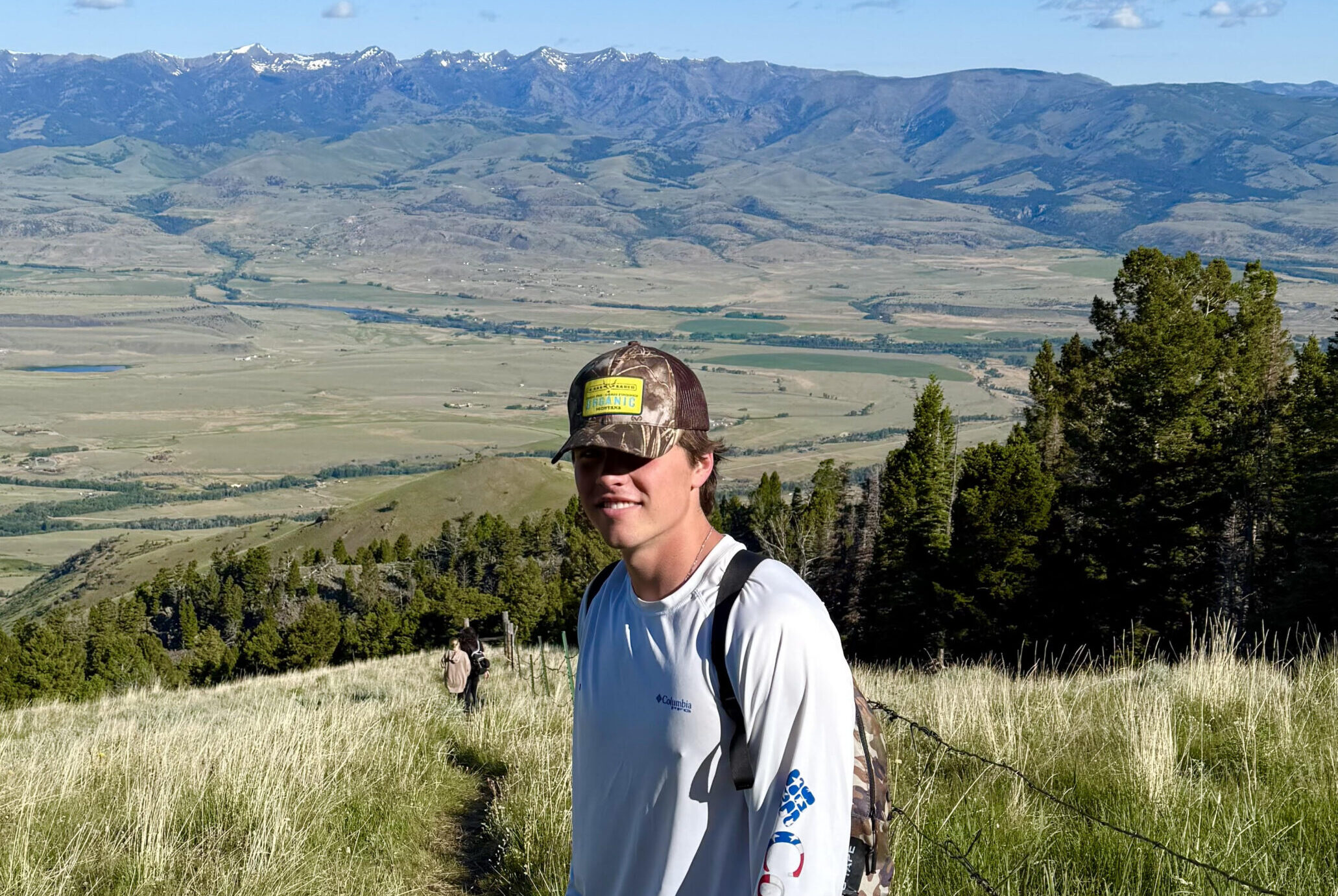 Young hiker in a camo cap and white long-sleeve shirt stands on a grassy ridge trail above a broad valley and snow-capped mountains.