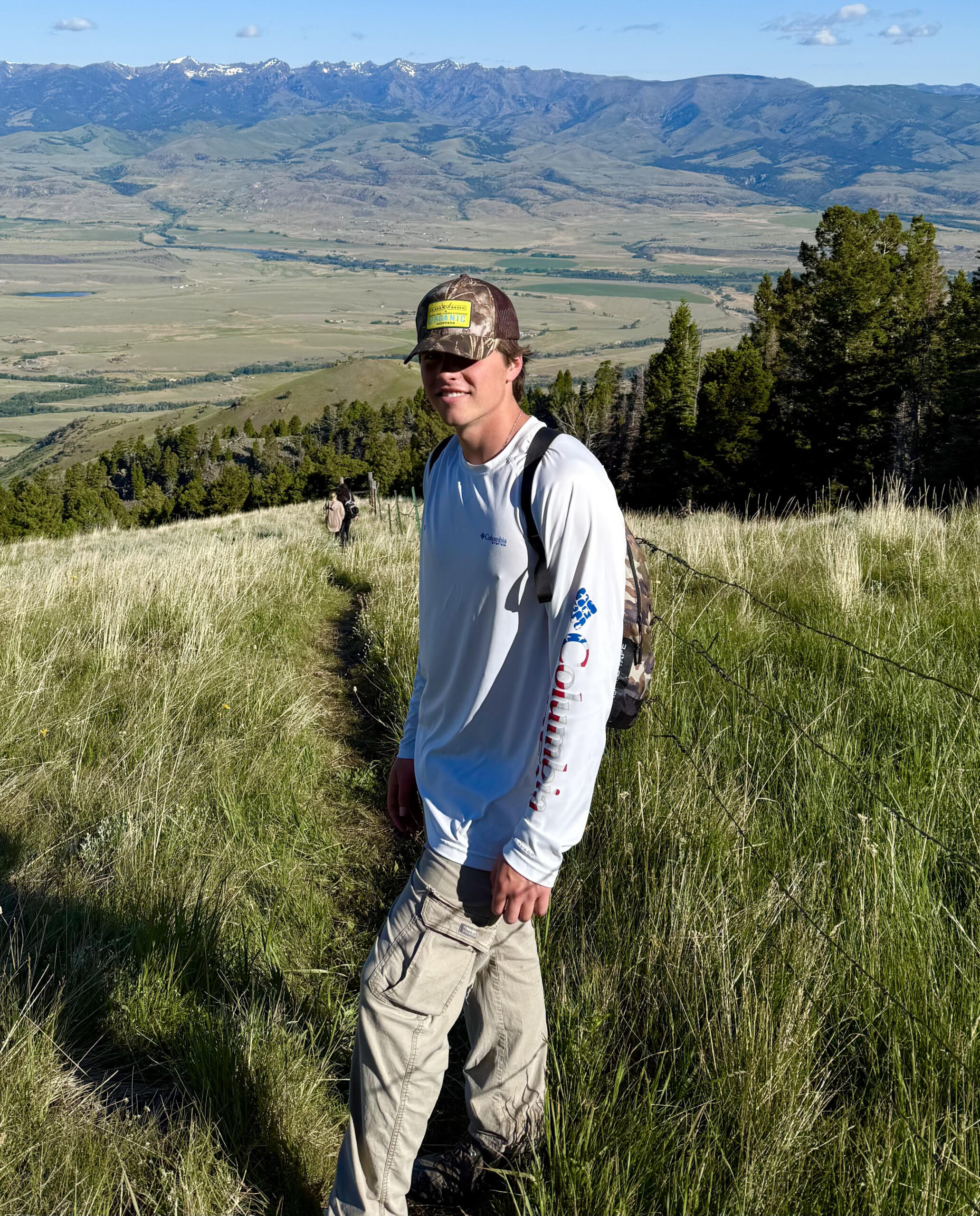 Young hiker in a camo cap and white long-sleeve shirt stands on a grassy ridge trail above a broad valley and snow-capped mountains.